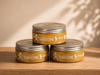 Three gold tins of Barbertime pomade stacked in a pyramid arrangement on a wooden surface with soft sunlight